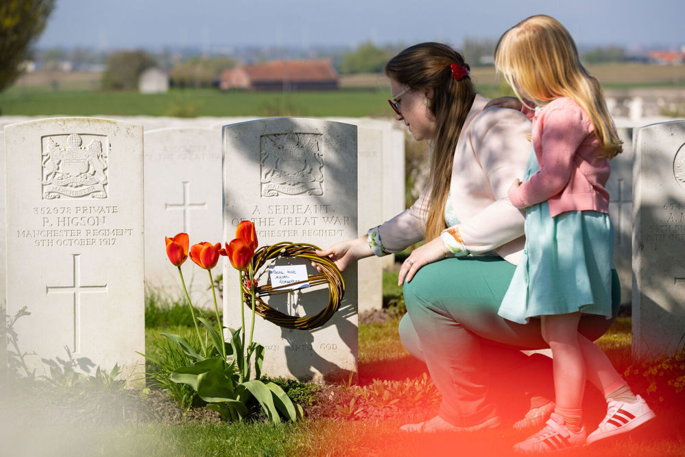 A mother lays a wreath on a CWGC headstone while her young daughter looks on.