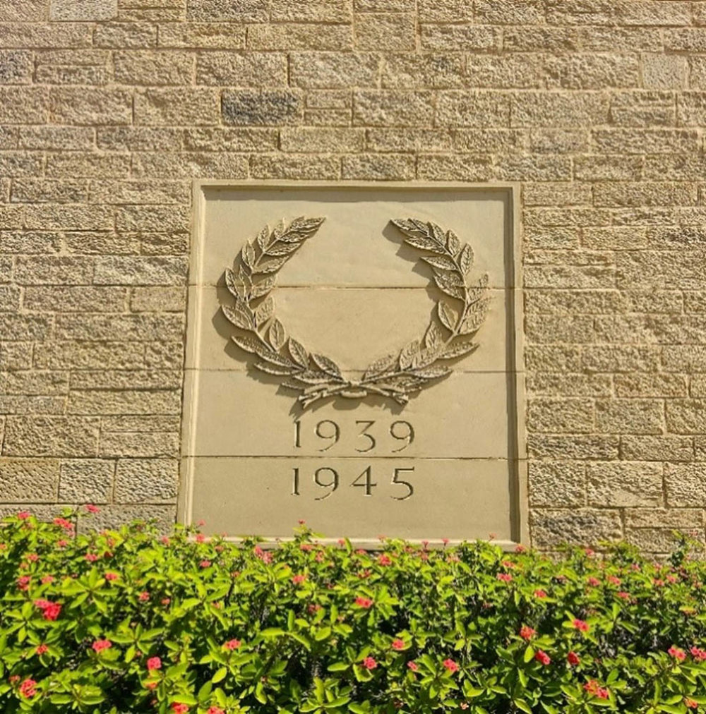 Laurel Wreath engraving on the wall at Delhi War Cemetery. Text reads 1939-1945. Below the wall carving sites a thick green bush with red flowers.