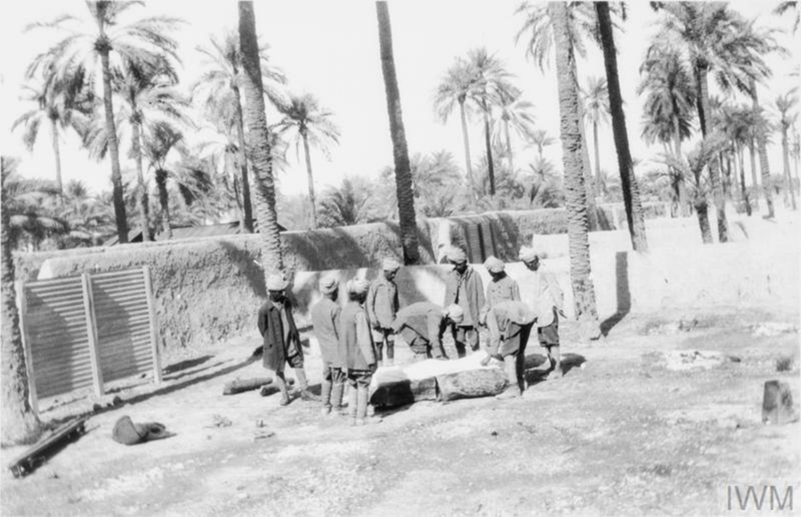 Four WW1 era Indian army soldiers preparing a bier for the cremation of a comrade underneath tall palm trees next to a low wall.