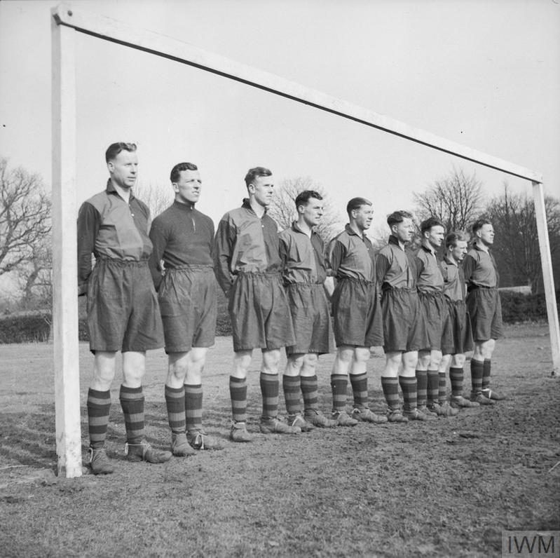 Men from Bolton Wanderers Football Club serving together with a battery of artillery (53rd Field Regiment, Royal Artillery, 42 Division, 11 Corps). THey are wearing half-and-half coloured football shirts and standing on the goal line between the posts.