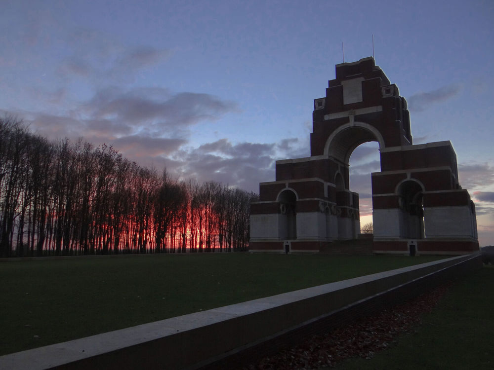 Thiepval Memorial at dusk on an autumn day. The low sun shines an orangey red glow behind autumnal trees.