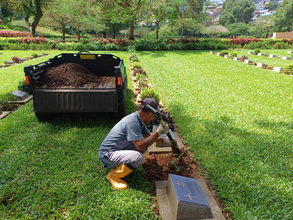 CWGC Gardener using a spade to pick up dirt and mulch for transferral to the compost cart behind him.