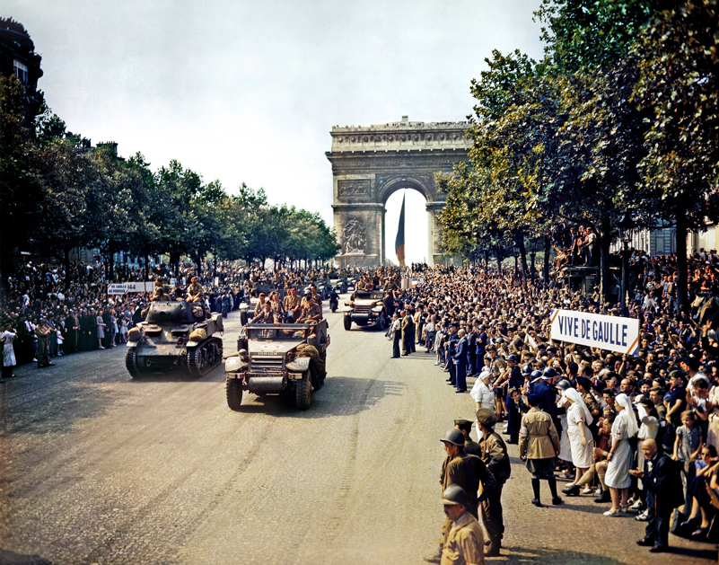Crowds of French Civilians line the streets as armoured vehicles parade victoriously up from the Arc de Triomphe.