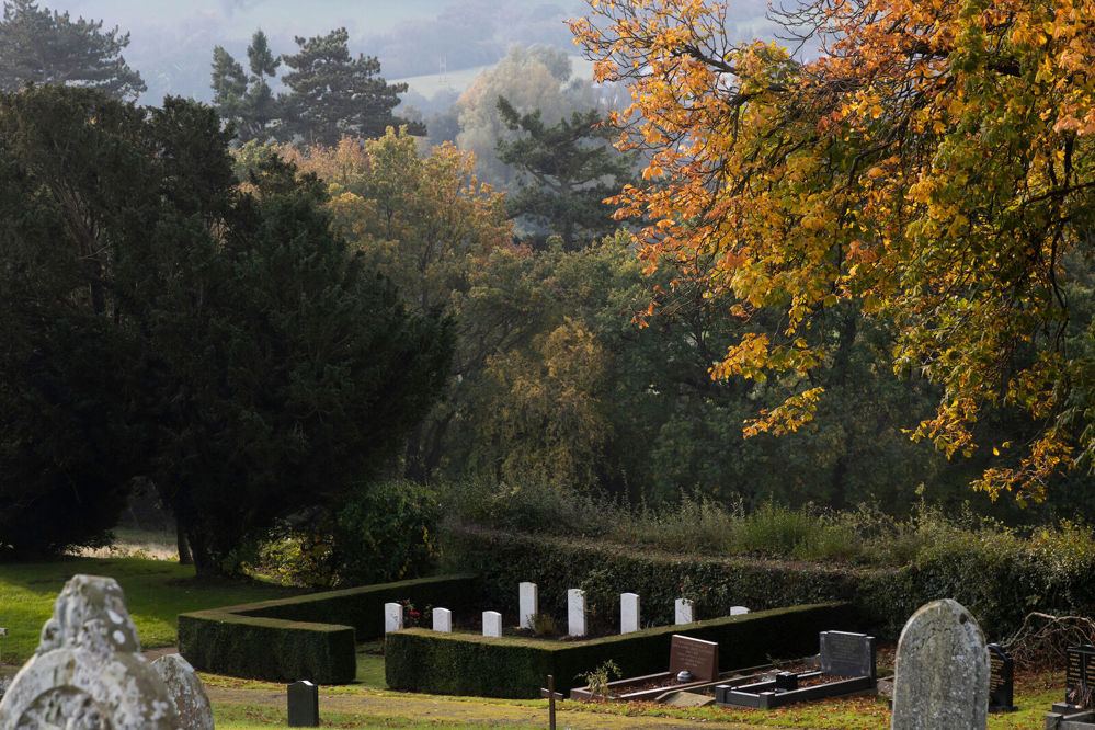 View of the Indian plot at Brecon Cemetery, showin gits thick, rectangular boundary hedge and white CWGC headstones. The trees around the cemetery are showing signs of the autumn season change. A number of old, weathered non-CWGC headstones are visible in the foreground.