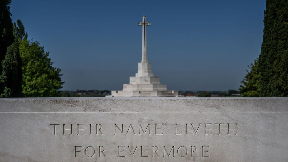 Stone of Remembrance with "Their Name Liveth for Evermore" inscription with CWGC Cross of Sacrifice visible in the background, featuring cuniform cross and bronze sword, set atop a stepped white stone plinth.