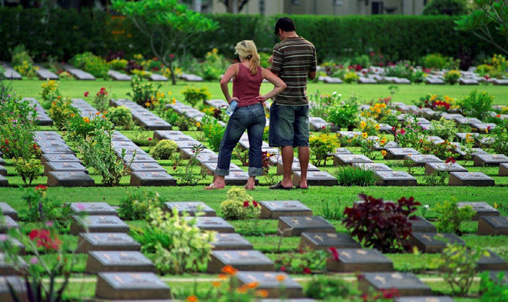 Two visitors examine bronze grave markers at Kanchanaburi War Cemetery, Thailand. The grave markers have been laid out in rows with a footpath bisecting the grave plots. A series of colourful plants sit in borders around the graves.