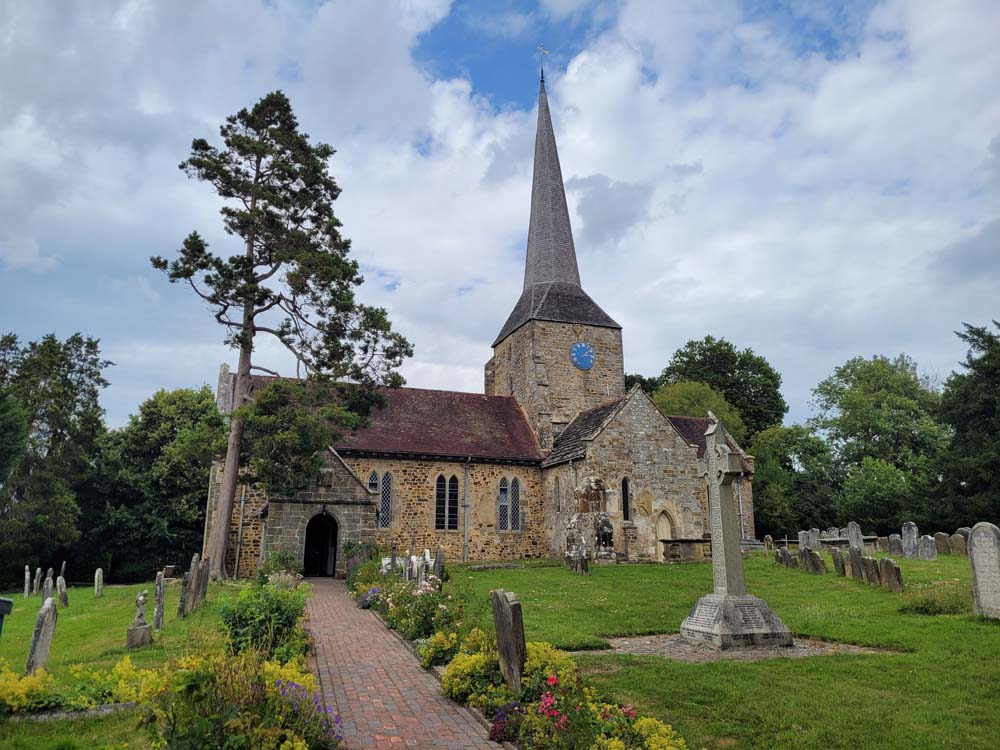 Church with central tall triangular spire. A well kept lawn with a few headstones can be seen in the churchyard alongside tall green trees in front of the church.