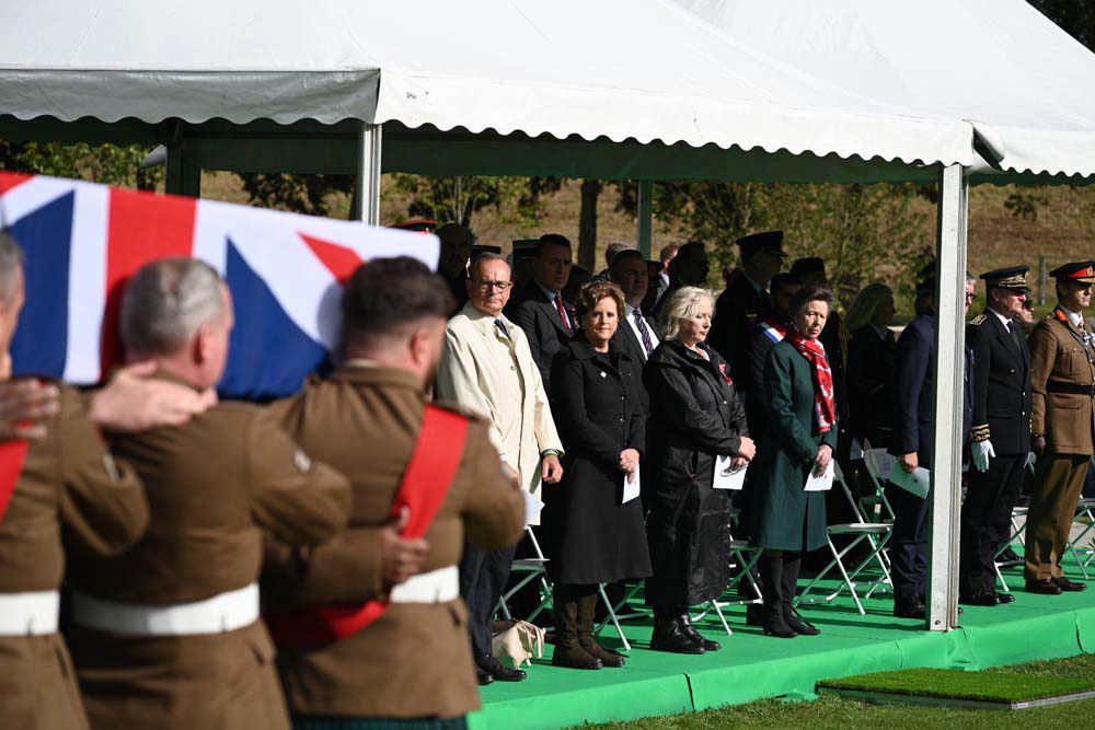 Princess Anne and several dignataries watch as a funeral procession carries a Union Jack-draped coffin past gazebo at Loos British Cemetery