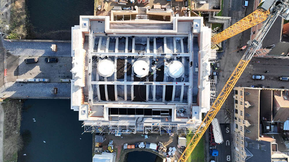 Aerial view of the Menin Gate Memorial showing its new eco roof under construction. Portions of turf have been laid in rectangular sections to make up the ribbed Memorial Roof. Three roud oculi sit in the central section but have been covered by a white covering. A yellow crane stretches over the scene.