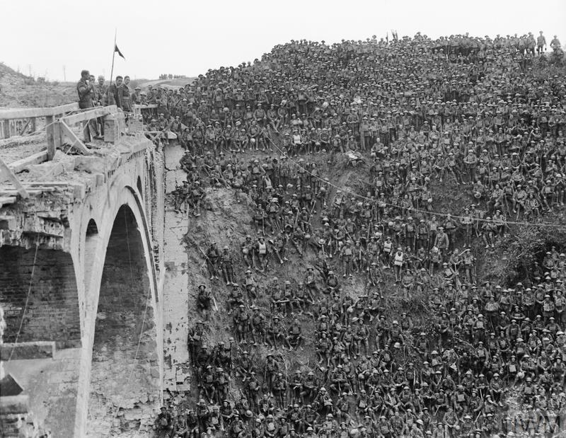 Thousands of British Infantry men sat on a canal cutting being addressed by their commanding officer. The CO is standing on a ruined bridge.