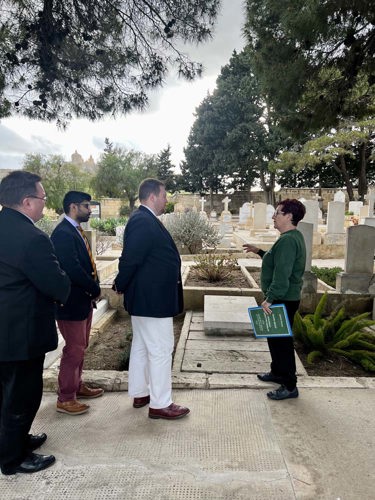 A short middle-aged woman in a green jumper gives a tlak to three gentlemen in a CWGC cemetery.