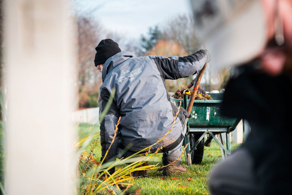 CWGC gardner kneels down next to a wheelbarrow while workin on plant borders. In the foreground you can see an out-of-focus close up of another gardener.