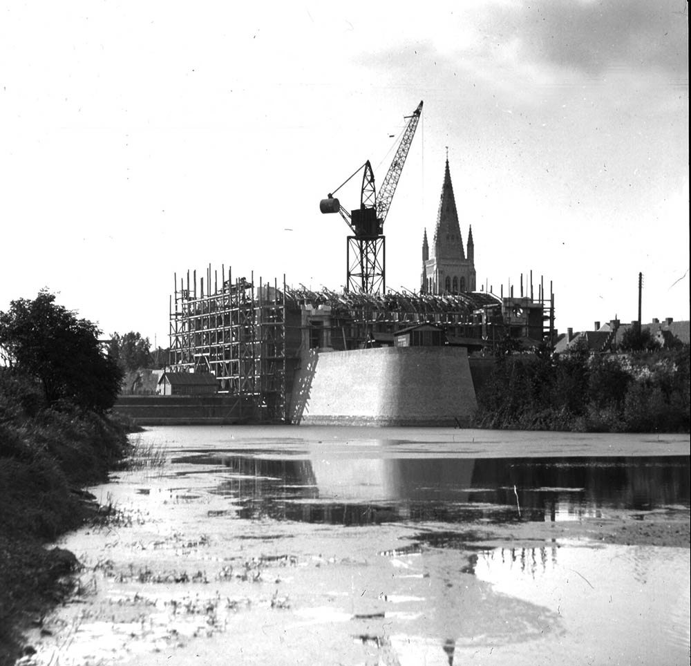 The Menin Gate Memorial under construction in the mid-1920s. A crane sits in the middle of the structure, rising from the old town ramparts. The main hall is taking shape, surrounded by a web of scaffolding. A church spire can be seen in the background.