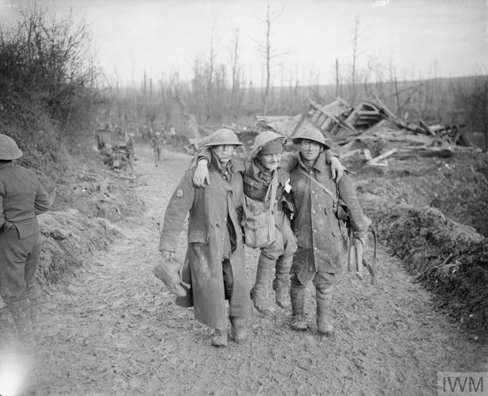 Two British soldiers carry a wounded comrade up a muddy road during the battle of the Somme. Wreckage and debris is visible in the background behind the travelling party.