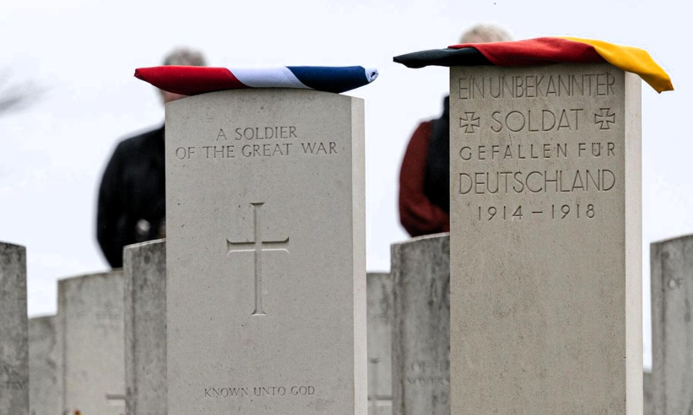 A CWGC British headstone next to a German first world war headstone. A folded Union Jack has been placed on top of the British headstone while a folded German flag sits on the German grave marker.