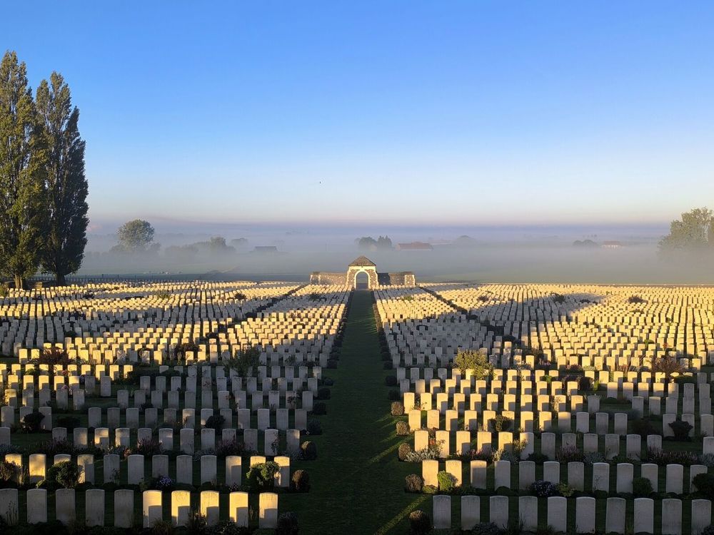 View of Tyne Cot Cemetery during a misty sun rise. Exstensive rows of white round-topped CWGC headstones stretch down into the distance towards the stone shelter building at the foot of a hill. The view behind the cemetery is obsucred by a thick misty blanket, gradually turning from grey to blue as it rises beneath the sky.