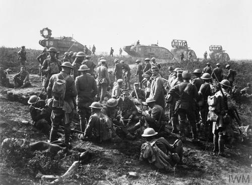 First World War American soldiers with Brodie helmets and full kit wait on a road side while British heavy tanks pass by in the background.