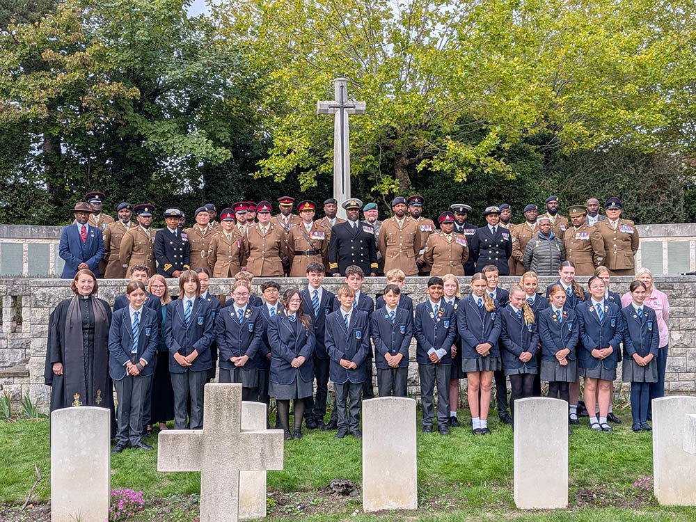 Two rows of people pose for the camera in Southampton Hollybrook Cemetery. The first row is two lines of schoolchildren in blue and dark grey unifroms, flanked by a priest and teachers. The back row is black, white, and Asian British servicemen from multiple military branches, plus other dignitaries.