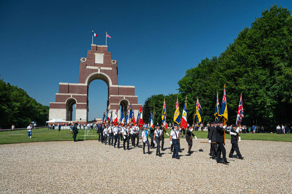 A parade of ex servicemen holding banners and flags marches along the gravel path in front of the Thiepval Memorial