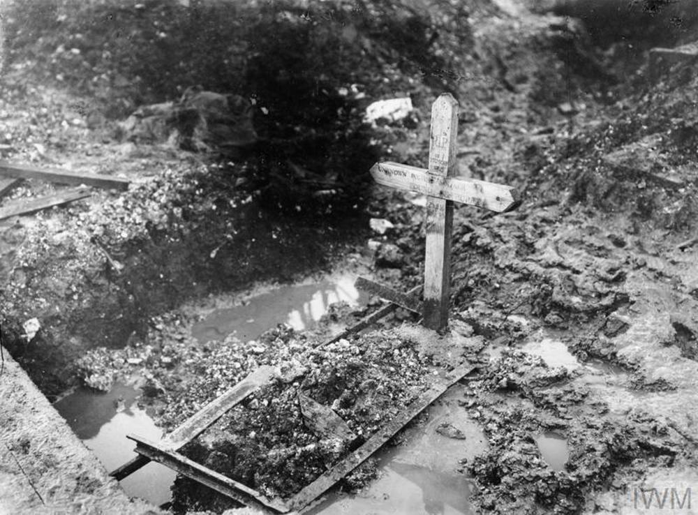 A temporary wooden cross grave marker marks the war grave of an unknown British soldier of the Somme. The grave itself is churned up mud bounded with simple wooden slats. To the left is a muddy shell puddle.