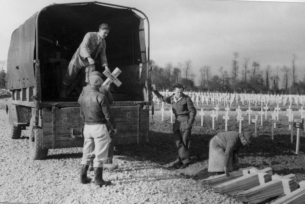 A man passess wooden grave markets to workers out the back of a WW2-era lorry. Stacks of wooden cross grave markets have been piled next to the road. In the background are visible rows of installed cross grave markets.