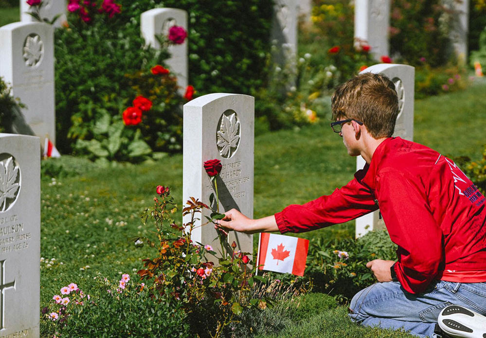A young boy in a red jumper places a red rose on the war grave of a Canadian soldier. A small Canadian flag has been placed in the grave border.