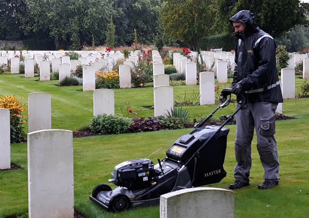 Man with a lawnmower mows the grass at Harrogate Stonefall Cemetery. Lots of white stone CWGC headstones are visible in the background.