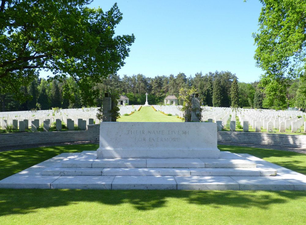 Stone of Remembrance at Becklingen War Cemetery. Behind the stone of remembrance is a stone gateway with the two posts, topped with round stone features, wrapped with plants. A wide grass drive leads to the Cross of Sacrifice in the background. On either side of the path site rows of white CWGC headstones.