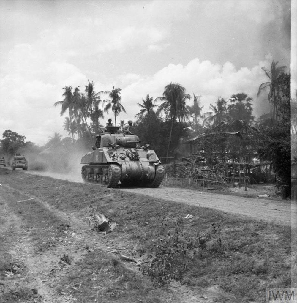 Indian Army sherman tanks advance along a jungle road in Burma. A variety of tropical trees are visible in the background while one side of the road has been cleared of shrub and trees.