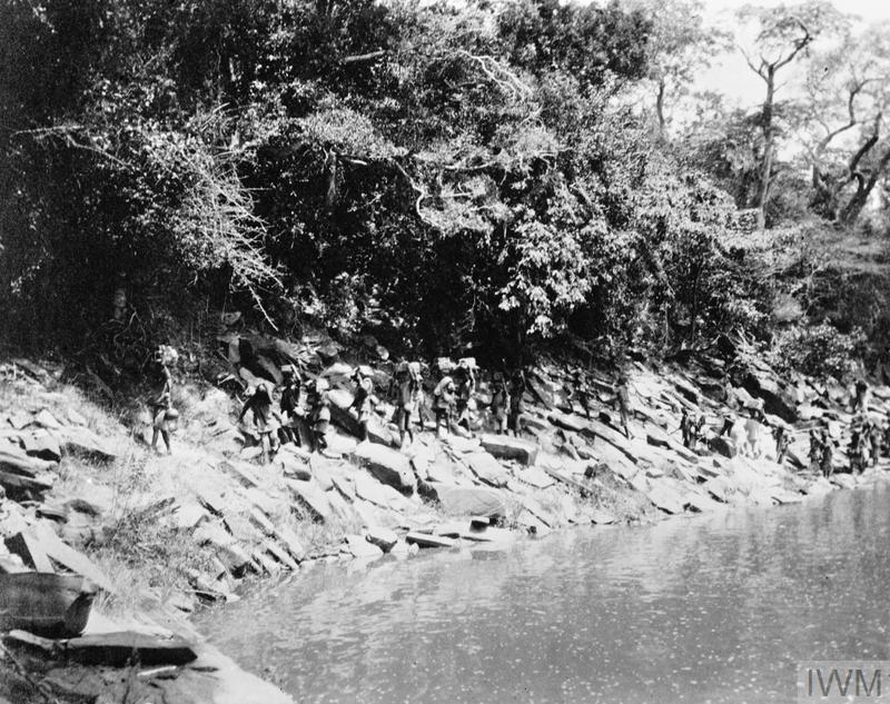 A column of WW1 era Nigerian soldiers in khaki fatigues move passed a river.