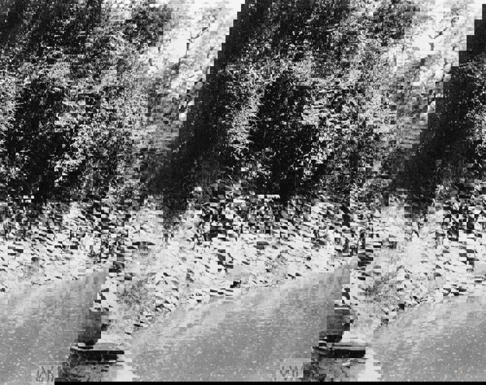 A column of WW1 era Nigerian soldiers in khaki fatigues move passed a river.