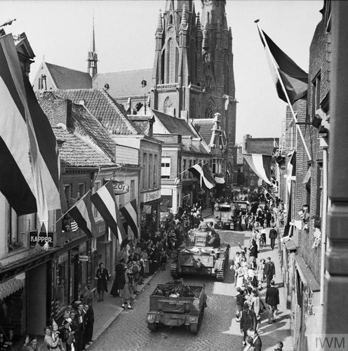 Dutch civilians swarm British tanks as they pass through a town street in Eidenhoven during Operation Market Garden. Many Dutch flags fly from the buildings.