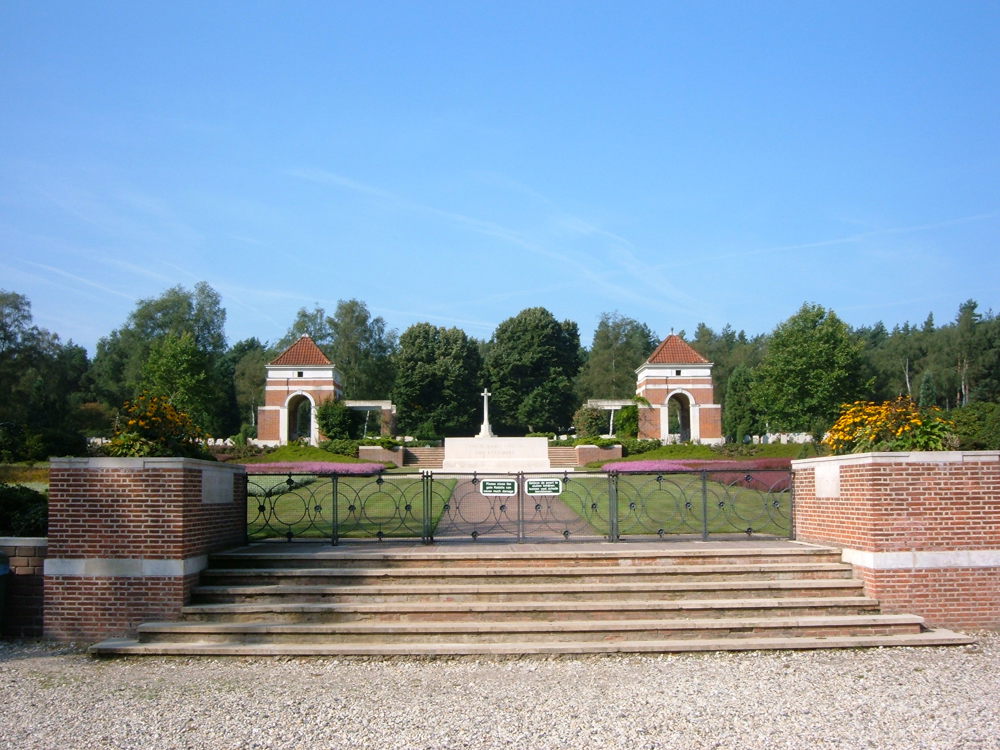View of Holten Canadian War Cemetery. A set of stone steps, flanked by two red brick planters topped with colourful flowers, lead up to black iron gates. The gates feature circular patterns in the ironwork. Beyond the gate lies the white Stone of Remembrance with the Cross of Sacrifice in the background. Two pointed, arched red brick towers flank either side of the cemetery plots.
