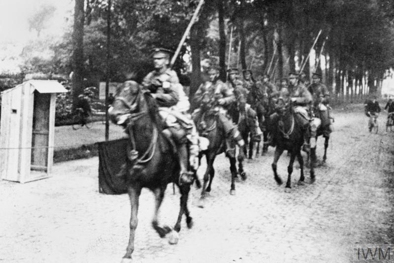 Horseman of the 9th Lancers arriving at Mons along a cobbled street under an avenue of tall trees.