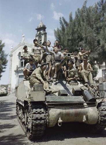 A group of sicilian children sat aboard a British Sherman tank. A picturesque church can be seen in the background as can fir trees.