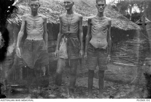 Three Australian PoWs pose in front of a low wicker hut. The three men are extremely malnourished with ribcages, shoulders, abd bone structure clearly visible. Shirtless, they are wearing tattered, dirty rags to cover themselves. They are barefoot.