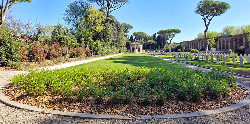 View of Rome War cemetery showing a low shot of the centralgrass lawn with the round Roman-esque entry building visible in the background. To the right, you can see the Antonine Walls lining the cemetery, while a wall of trees, hedges, and other plantlife is visible on the left flank.