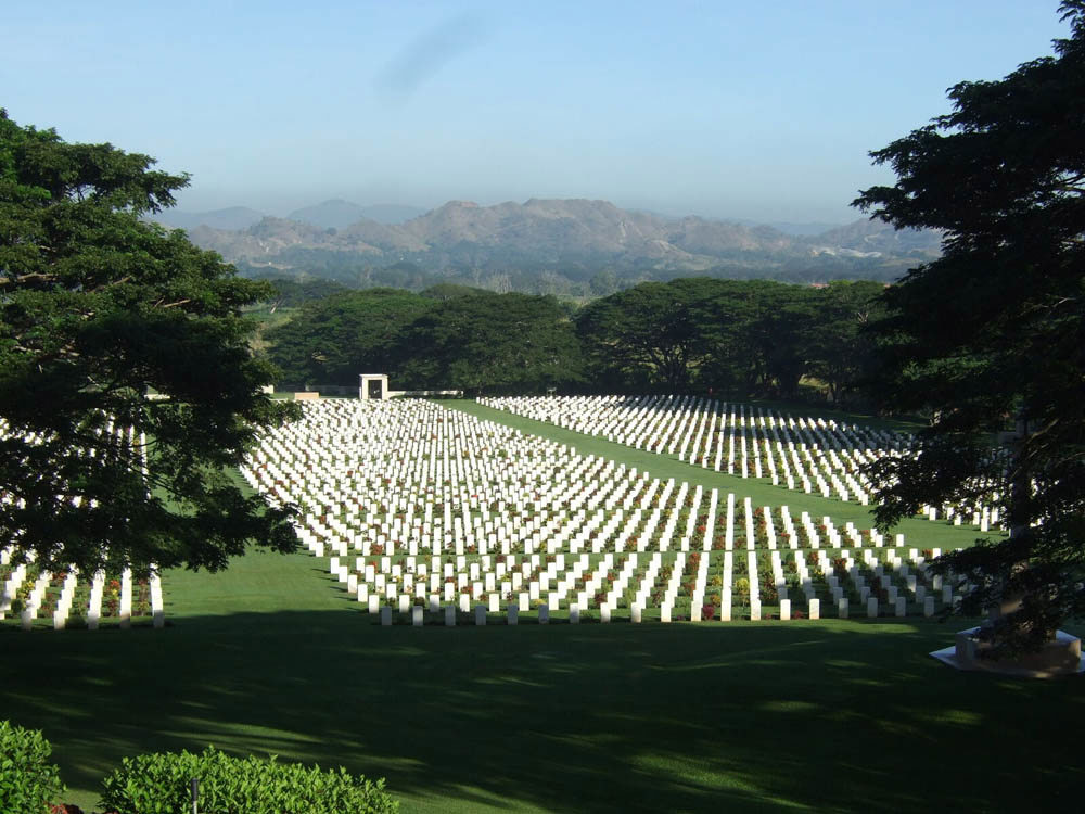 View of Ports Moresby War Cemetery from a top a hill showing the wide cemetery lawns and paths and multiple rows of white CWGC headstones.