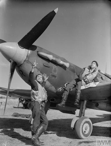 Two West Indian pilots relaxing on the wing and engine cowling of a Spitfire aircraft.