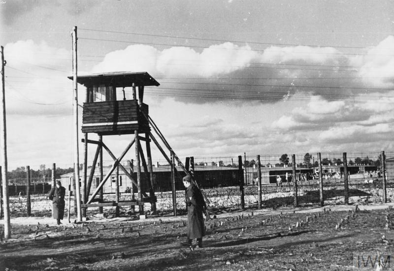 View of a watchtower at Stalag Luft III. Barbed wire fences can be seen surrounding wooden huts while a guard stands sentry.