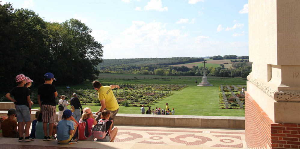 A CWGC Guide wearing a yellow polo shirt gestures towards the horizon while talking to a group of schoolchildren on the steps of the Thiepval Memorial.  The group is facing the Thiepval Anglo-French Cemetery, featuring rows of headstones and grave markers along a central wide lawn, topped with the Cross of Sarifice.