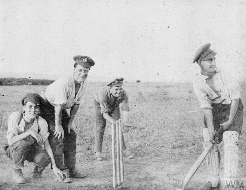 Four WW1 British soldiers pose in front of a wicket and stumps. They are in more casual clothing but are still wearing their peaked military caps. One is holding a cricket bat, looking to receive a ball.