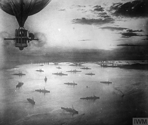 Black and white image of naval ships lying in anchor of the Firth of Forth. The Forth Rail Bridge can be seen in the background. The ships are silhouetted against the sea.