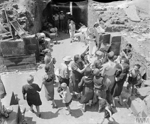 A crowd of civilians surround an RAF servicemen who is doling out food parcels amongst the rubble of some damaged Sicilian buildings.
