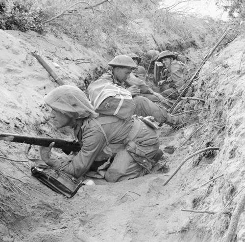 British WW2 soldiers sat in a shallow trench. The soldiers are carrying rifles and wearing helmets.