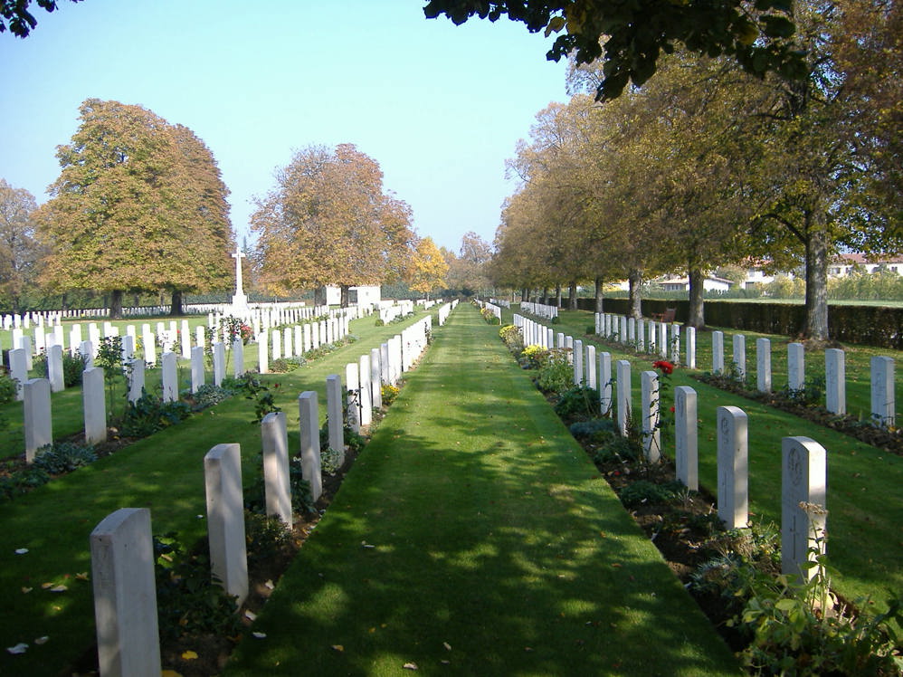 Rows of headstones at Faenza War Cemetery. Tall trees, with autumnal colours, stand over the headstones and green lawn.