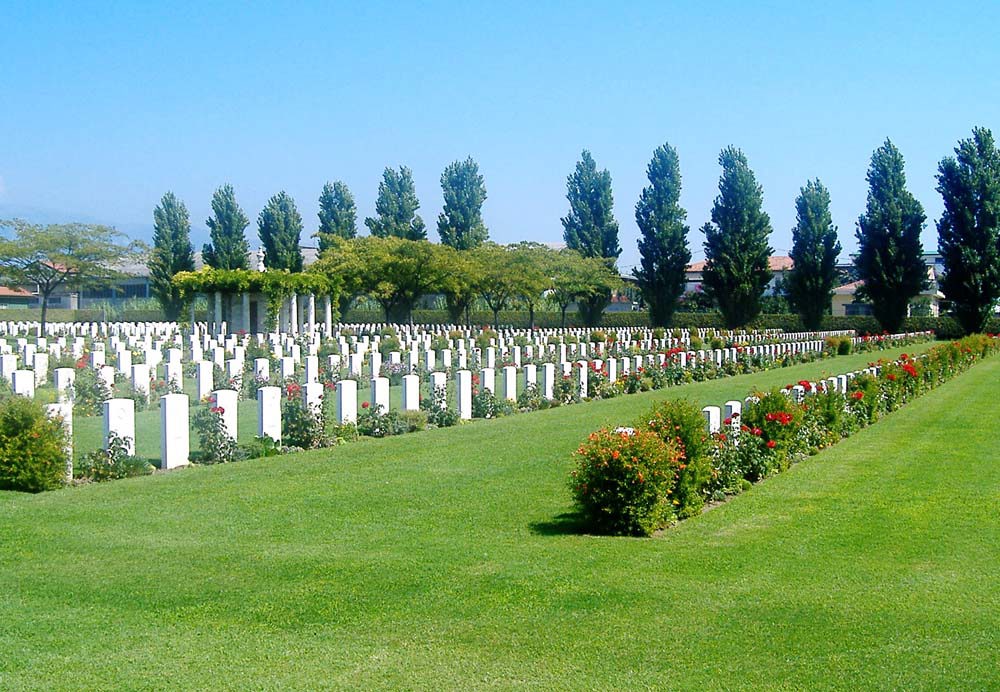 Rows of headstones at Salerno War Cemetery. A rectangular shelter building topped with greenery can be seen in the background. The white headtones sit atop a neatly mown green lawn. A row of tall trees can be seen in the background.