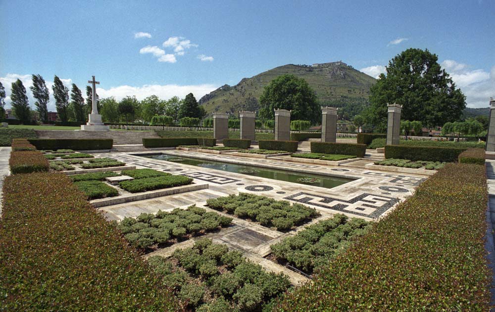 Cassino War Cemetery's central architectural feature, showing a reflective pool surrounded by Roman-inspired mosaic tiling and neatly trimmed architectural features. Monte Cassino itself is visible in the background under a clear blue sky.