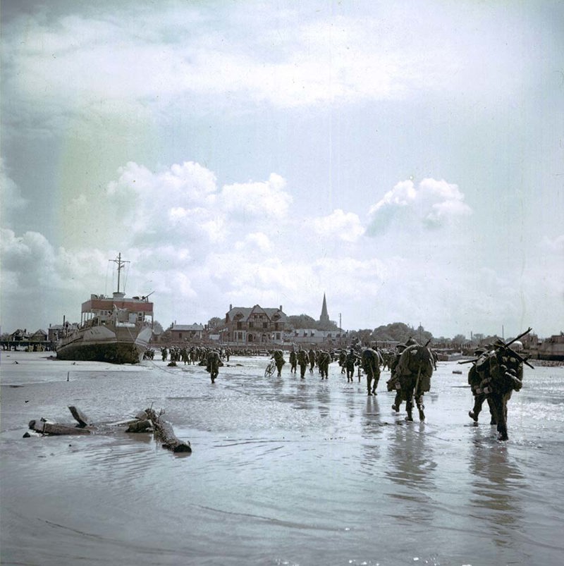 Canadian soldiers marching up Juno Beach. A landing craft can be seen in the background as can an array of buildings on the beachfront.